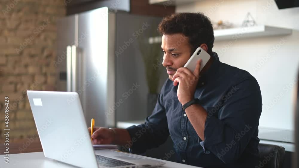 Young dark-haired indian male entrepreneur using laptop in the kitchen at home, talking on the phone and takes notes, multitasking senior good looking man working remotely from home Young dark-haired indian male entrepreneur using laptop in the kitchen at home, talking on the phone and takes notes, multitasking senior good looking man working remotely from home