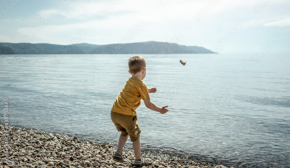 Cute little boy is throwing stones into the water on the shore of a large pond. Concept of summer children's leisure, outdoor activities, vacations and travel