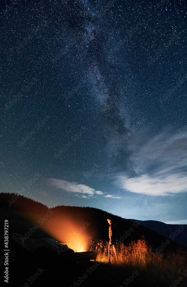 Man hiker looking at bright bonfire while standing under blue night sky with stars and Milky Way. Starry sky over grassy hill with male traveler near campfire. Concept of night camping and astronomy.