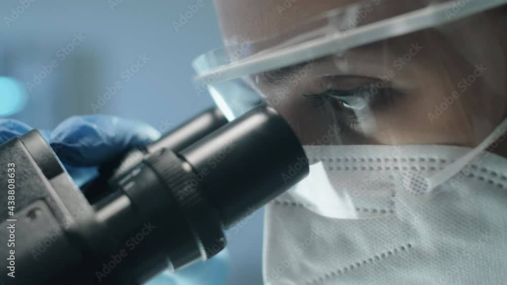 Close up shot of female scientist in protective uniform, mask and glasses looking through microscope while doing lab research during covid- outbreak Close up shot of female scientist in protective uniform, mask and glasses looking through microscope while doing lab research during covid- outbreak