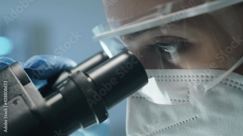 Close up shot of female scientist in protective uniform, mask and glasses looking through microscope while doing lab research during covid- outbreak photo