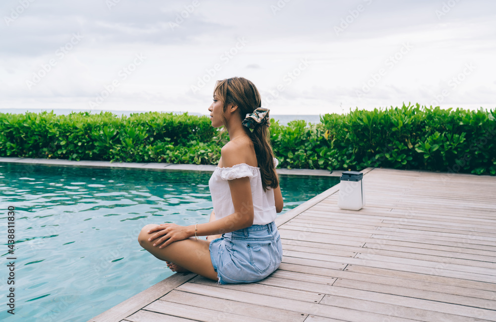 Calm young ethnic female sitting on pool wooden deck with crossed legs