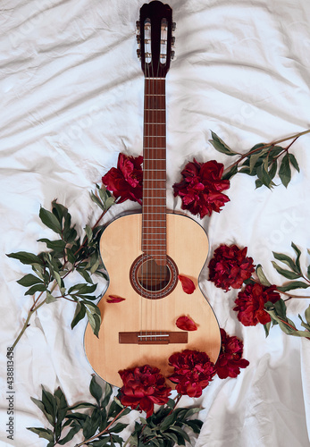 An acoustic wooden guitar lies on a white sheet surrounded by red peonies.