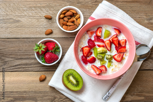 oatmeal porridge with slices of kiwi, strawberries, almonds in pink bowl, spoon, napkin with red stripes on wooden background Vegan food concept Healthy breakfast