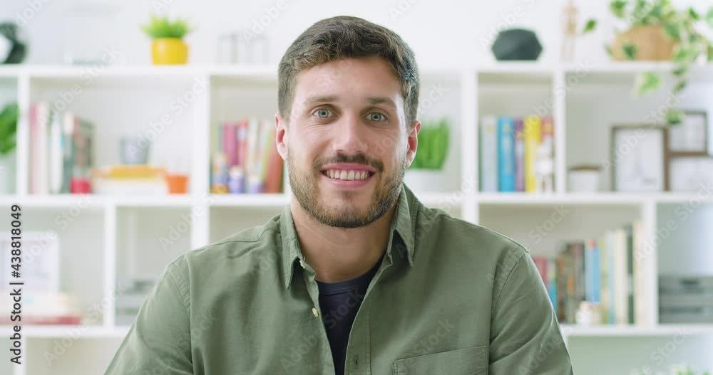 Slow motion of handsome smiling confident young caucasian man looking at camera smiling in living room or home office. Happy millennial authentic real male close up front portrait. Slow motion of handsome smiling confident young caucasian man looking at camera smiling in living room or home office. Happy millennial authentic real male close up front portrait.