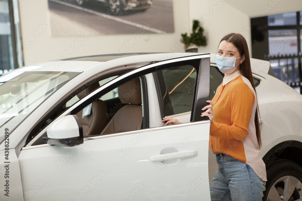 Young female customer wearing medical face mask at car dealership, choosing new automobile to buy