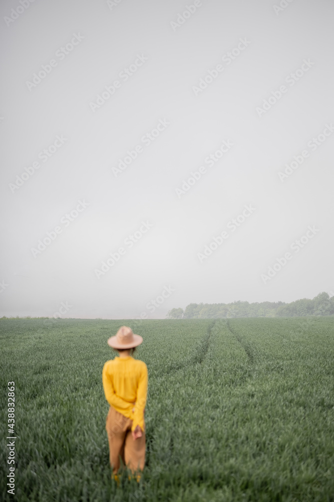 Woman in hat walking on green wheat field during foggy weather. Countryside and quiet living. Back view. Person is out of focus.