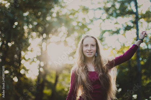 A portrait of a beautiful smiling woman with long blond hair in a purple blouse standing in the park, raising her left hand up. Close up photo of a girl with back sunlight on the nature