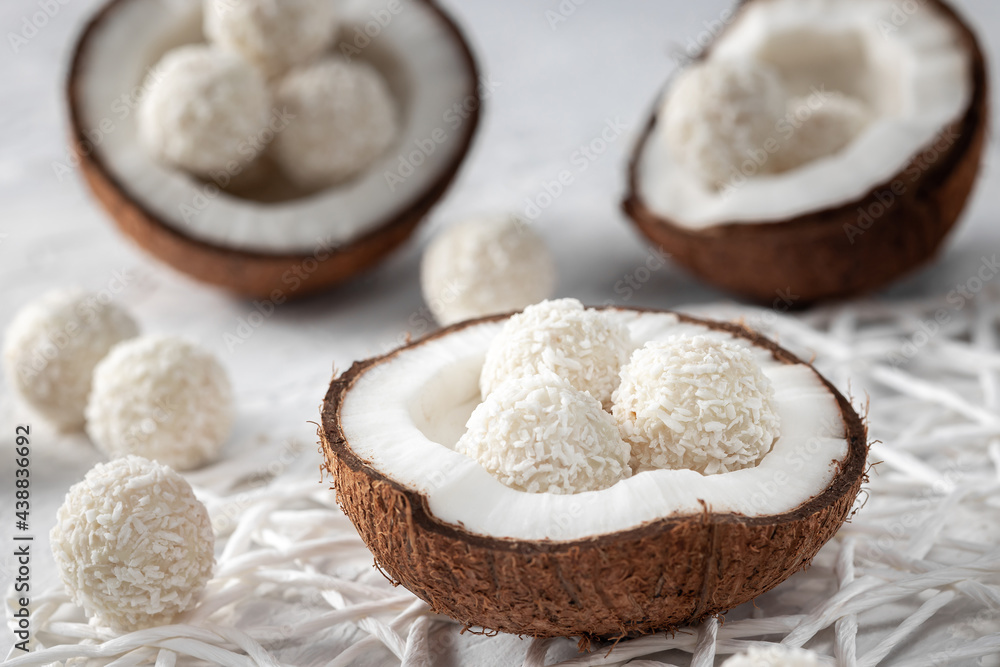 Portions of white chocolate coconut candy balls in the bowl, raw cracked coconut on the background, selective focus Portions of white chocolate coconut candy balls in the bowl, raw cracked coconut on the background, selective focus
