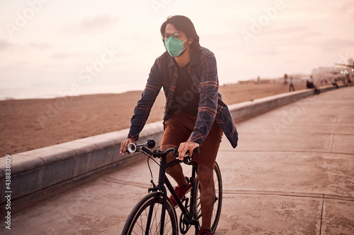 Fototapeta Naklejka Na Ścianę i Meble -  Hombre Joven latino maneja bicicleta durante un bello atardecer.. Joven feliz  con mascarilla en la playa