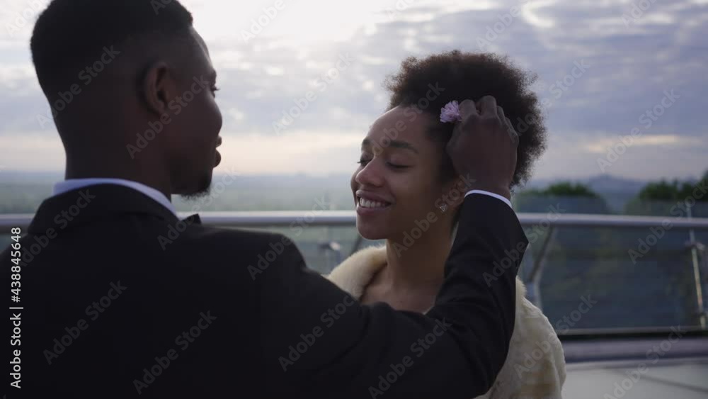 Portrait of smiling happy African American bride looking with love at groom putting flower in hair in slow motion. Positive confident young woman getting married with affectionate man on bridge Portrait of smiling happy African American bride looking with love at groom putting flower in hair in slow motion. Positive confident young woman getting married with affectionate man on bridge