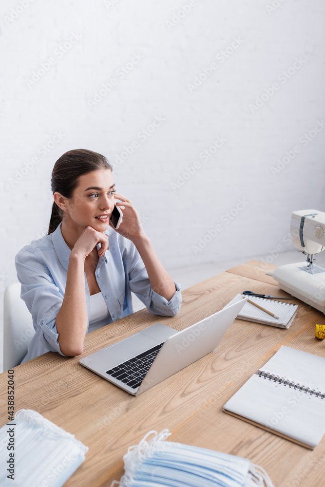 Smiling seamstress talking on smartphone near laptop and medical masks Smiling seamstress talking on smartphone near laptop and medical masks