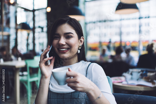 Joyful woman with cup talking on smartphone in cafe