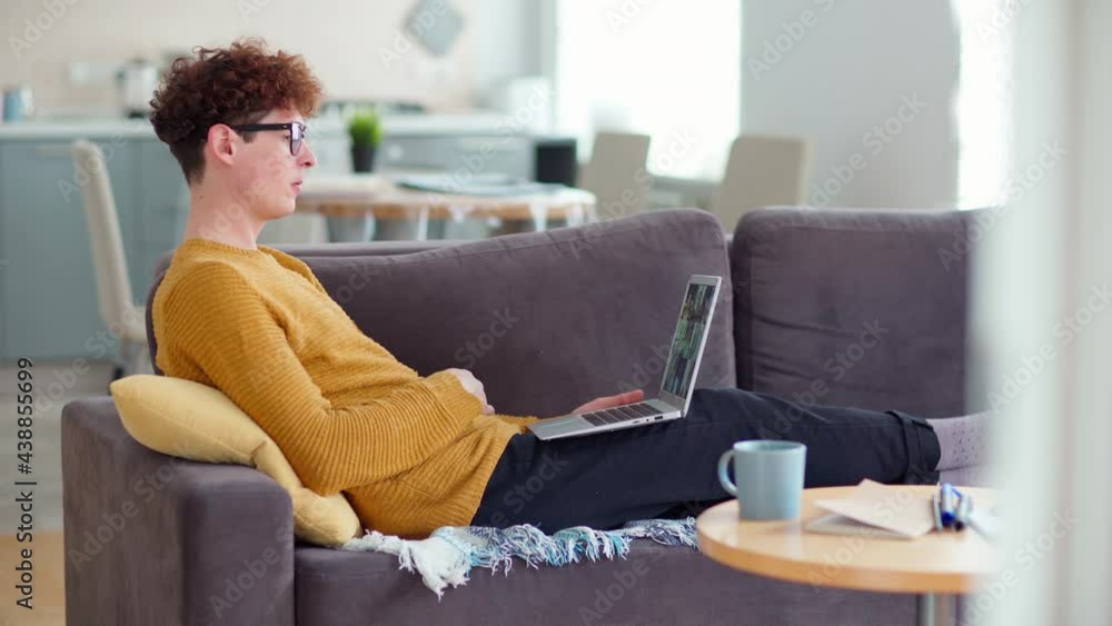 Side view tracking shot of young man in eyeglasses watching video conference on laptop lying on sofa. Male listening to group of colleagues while working remotely from home Side view tracking shot of young man in eyeglasses watching video conference on laptop lying on sofa. Male listening to group of colleagues while working remotely from home