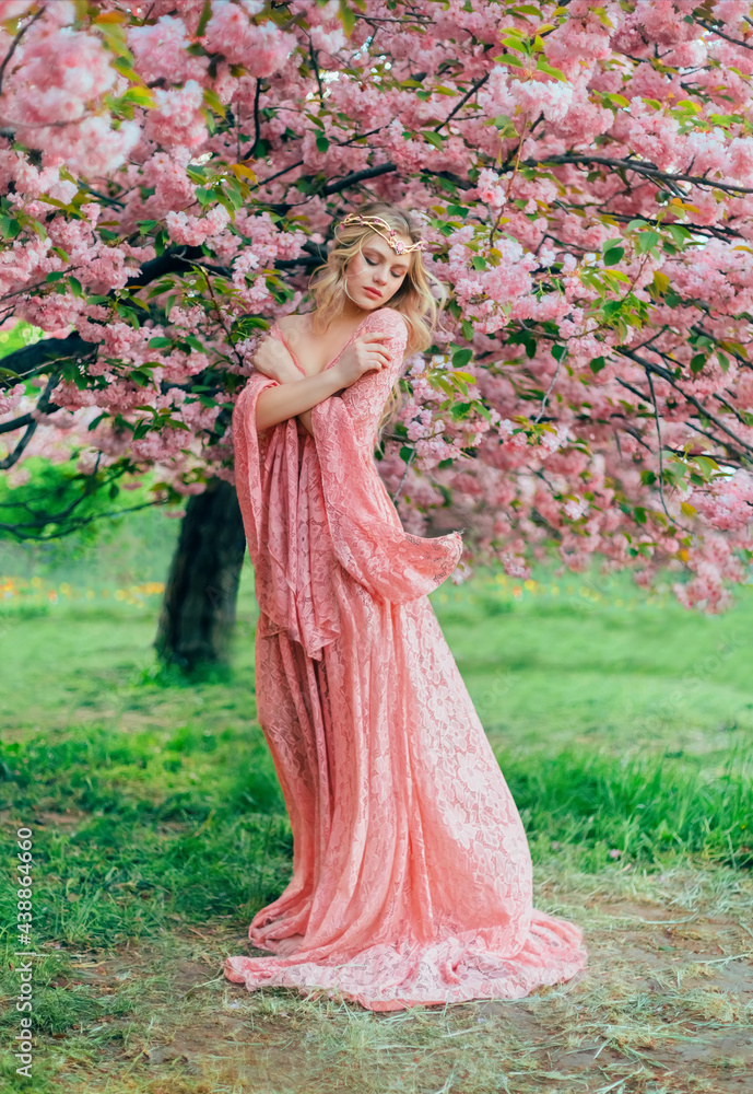 Fantasy happy woman princess walks in spring blooming garden hugs herself by shoulders. Pink flowers sakura tree summer nature. long lace dress wide sleeves flies in wind motion. Blonde girl queen.