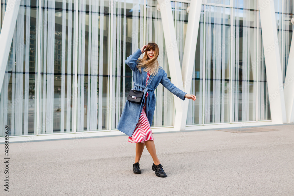 Happy brunette woman in blue coat and red dress walking along modern street