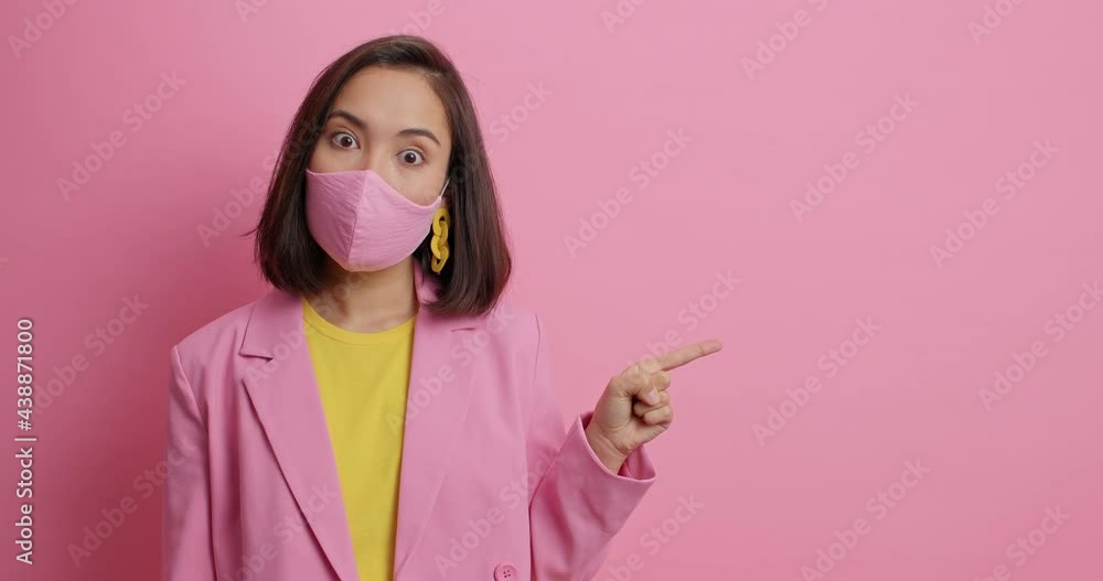 Covid 19 virus and epidemic concept. Surprised Asian woman wears protective mask to prevent contagious disease dressed in elegant jacket isolated over pink background shows copy space for promo Covid 19 virus and epidemic concept. Surprised Asian woman wears protective mask to prevent contagious disease dressed in elegant jacket isolated over pink background shows copy space for promo
