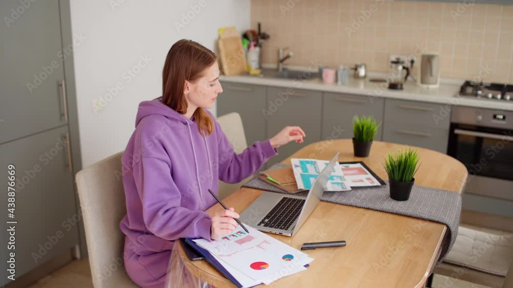 Young female marketing professional working from home in domestic kitchen. Woman examining papers with web interface templates and market research, browsing laptop and thinking, side view Young female marketing professional working from home in domestic kitchen. Woman examining papers with web interface templates and market research, browsing laptop and thinking, side view