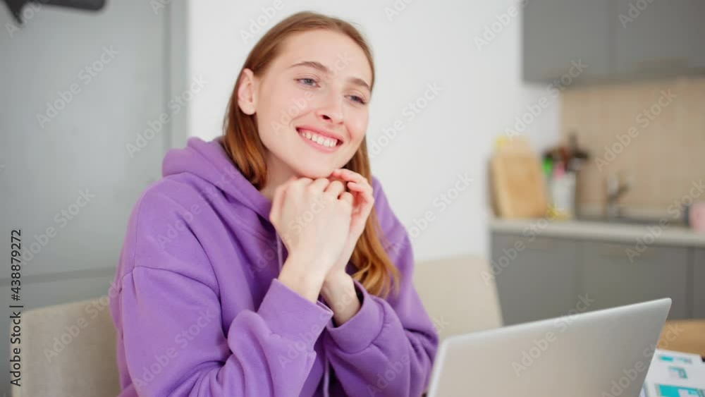 Beautiful young woman looking at camera and smiling happily while working on laptop at home. Happy female posing and day dreaming sitting at table with open laptop in front of her Beautiful young woman looking at camera and smiling happily while working on laptop at home. Happy female posing and day dreaming sitting at table with open laptop in front of her