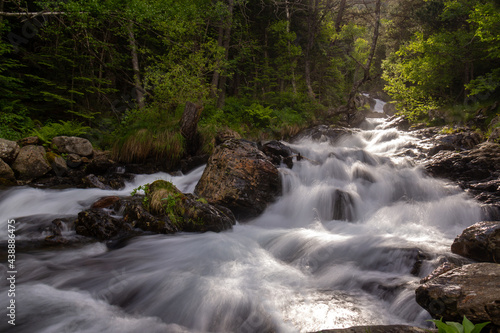 small river runs from the mountain through the stones and a colorful forest under the sunlight and along a path.