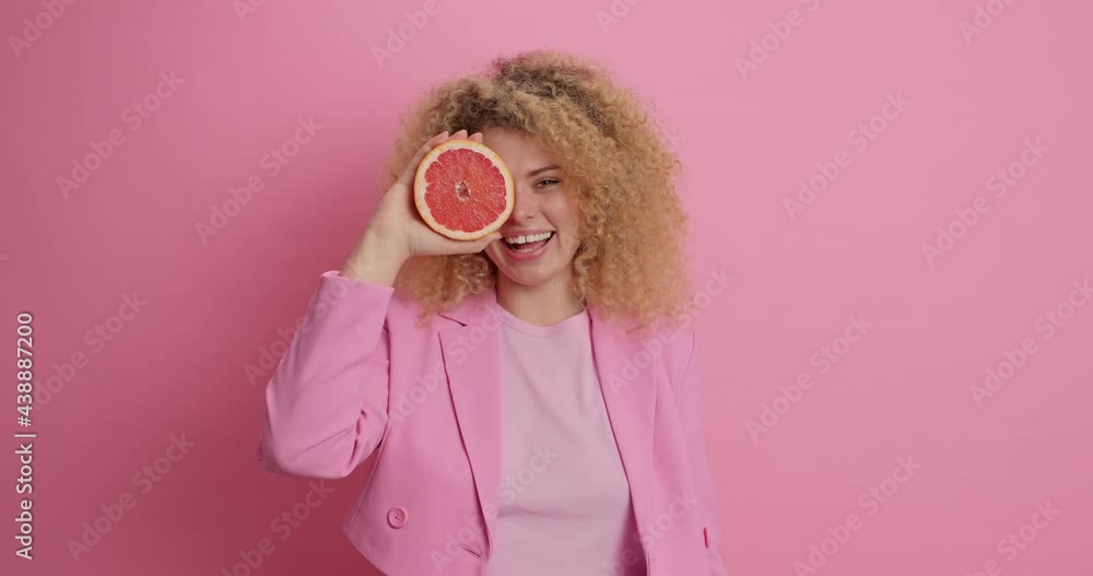 Healthy diet refreshing food full of vitamins. Glad European woman with curly hair covers eye with red grapefruit likes citrus fruit dressed in stylish clothes isolated over pink background. Healthy diet refreshing food full of vitamins. Glad European woman with curly hair covers eye with red grapefruit likes citrus fruit dressed in stylish clothes isolated over pink background.