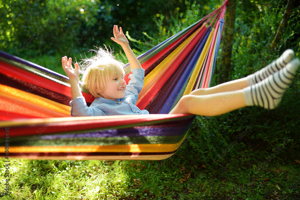 Cute little blond caucasian boy having fun with multicolored hammock in backyard or outdoor playground. Summer active leisure for kids. Child on hammock. Activities for children outdoors Cute little blond caucasian boy having fun with multicolored hammock in backyard or outdoor playground. Summer active leisure for kids. Child on hammock. Activities for children outdoors