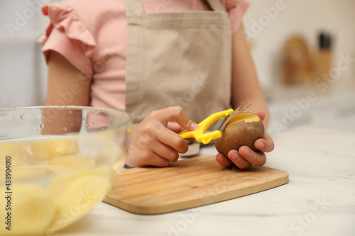 Little girl peeling potato at table in kitchen, closeup. Preparing vegetable