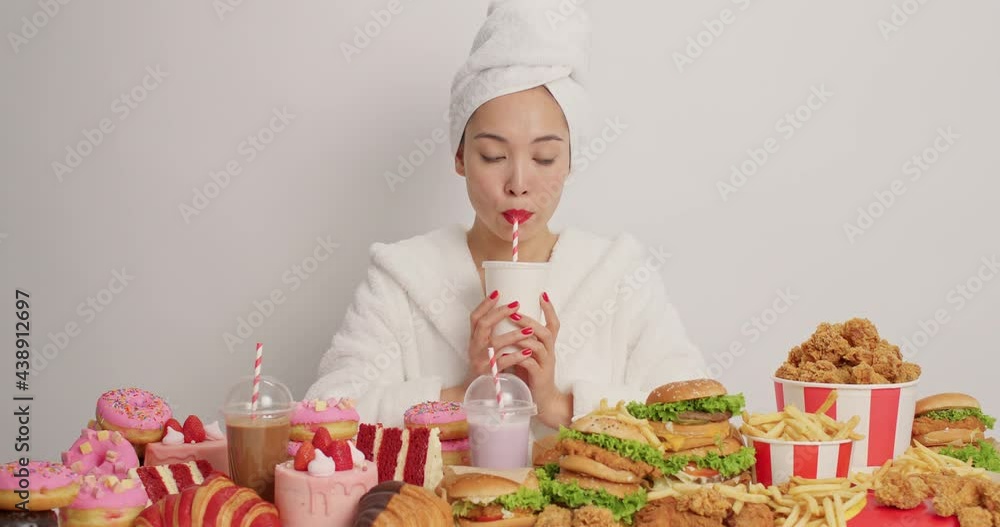 Surprised puzzled Asian woman looks at various junk food feels temptation to eat everything at same time drinks fizzy drink wears red lipstick bathrobe and towel over head isolated over white wall Surprised puzzled Asian woman looks at various junk food feels temptation to eat everything at same time drinks fizzy drink wears red lipstick bathrobe and towel over head isolated over white wall