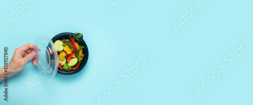 A female hand holds an open plastic disposable plate with vegetable salad on a blue background. Top view, flat lay. Banner