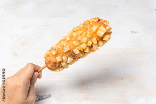Hand holding Korean street food corn dog is deep fried crusted flour stuffed with sausage and cheese on wooden skewer.