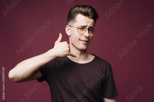 Young caucasian man wearing casual t-shirt and glasses standing over isolated crimson background smiling doing talking on the telephone gesture. Call me.