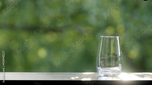 Empty clean glass on the brown organic wooden table with green morning summer or spring leaves of trees bokeh background. Sun's rays glare on a glass beaker