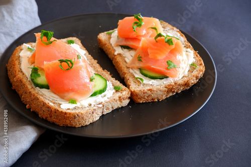 Toast with red fish, curd cheese and cucumber on a black plate.