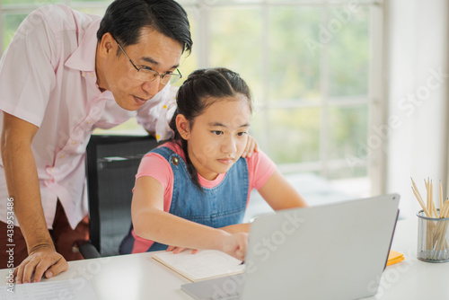 Young Asian love family daughter learning online use computer notebook and father help to teach in living room at home which smiling and felling happy. live at home concept in COVID-19 situation