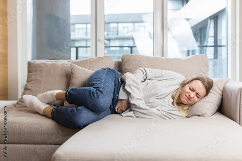 young woman sitting or lying on sofa or couch at home with severe abdominal pain. Female on bed holds her stomach with premenstrual, menstrual or poisoning. blonde girl suffers from abdominal cramps 