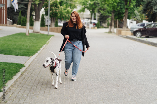 View of young Caucasian female cynologist walking at city urbanity during morning time for training dalmatian dog, plus size female spending leisure with pet best friend enjoying weekend at street