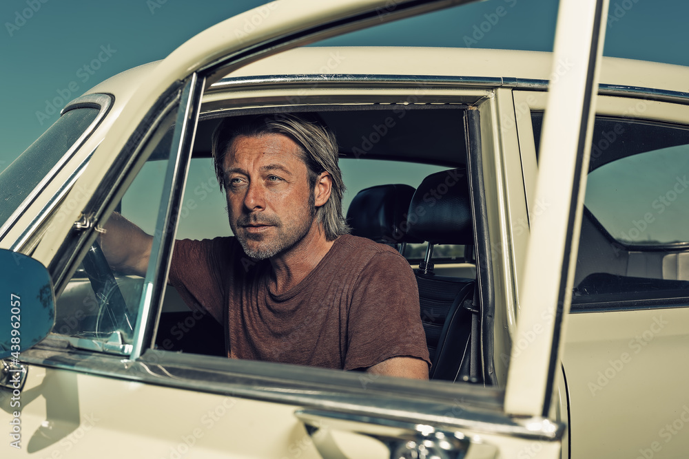 Blond man in t-shirt sitting in classic car with open door. Blond man in t-shirt sitting in classic car with open door.