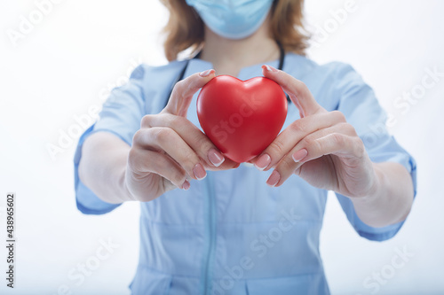 Close up of female doctor hands holding the heart. Red heart showing by woman doctor wearing blue medical face mask and lab coat with a stethoscope. Cardiology, healthcare, and medicine concept.