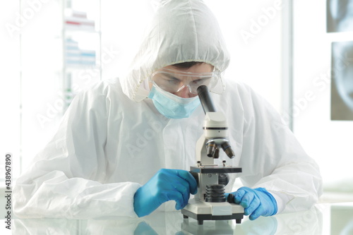 Doctor in a protective medical suit wearing a mask wearing blue gloves in a laboratory looks through a microscope
