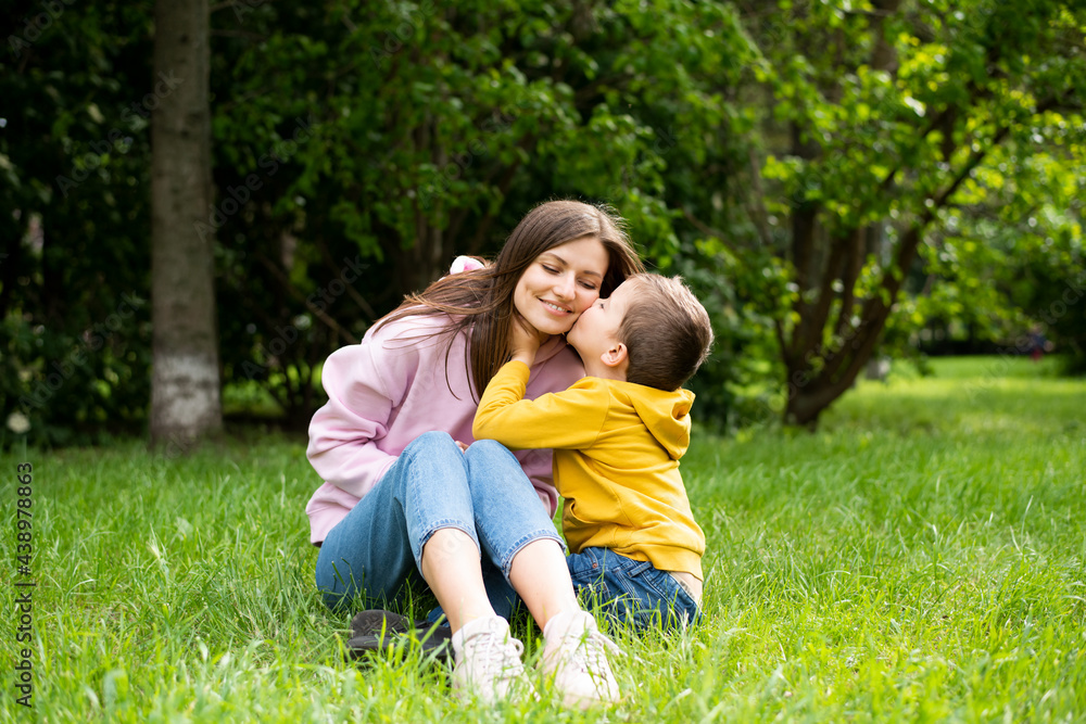 Young woman mom sits on green grass in summer park with boy son. Son kisses mom's cheek. Young woman mom sits on green grass in summer park with boy son. Son kisses mom's cheek.