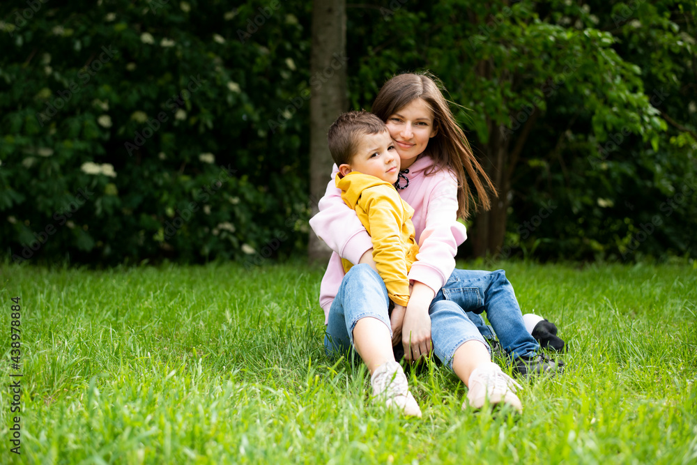 Young woman mom is sitting on green grass in a summer park with a boy son. Mom hugs her boy son. Young woman mom is sitting on green grass in a summer park with a boy son. Mom hugs her boy son.