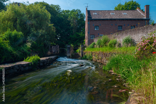 Long exposure of a water rapid of the Nekummer watermill in the Jeker river in the valley with blooming wild flowers and a old cottage just outside Maastricht