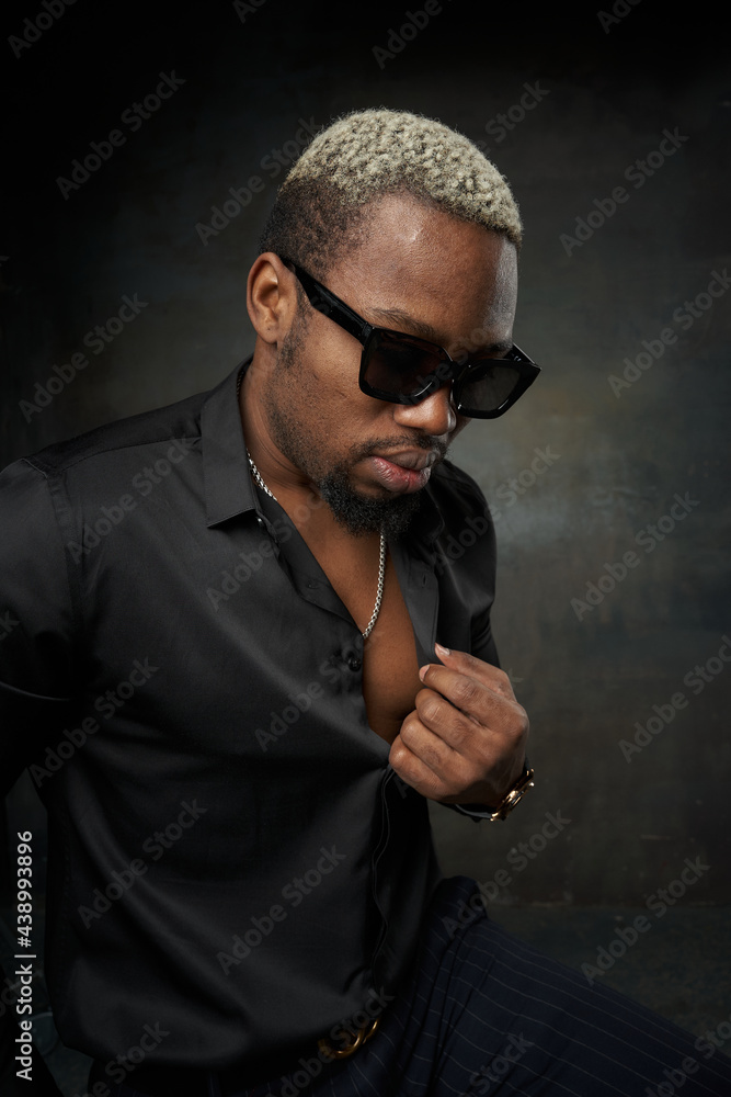 Close-up face studio portrait of young stylish glamor handsome african man with short white hair in black silk shirt and sunglasses posing against grey concrete wall