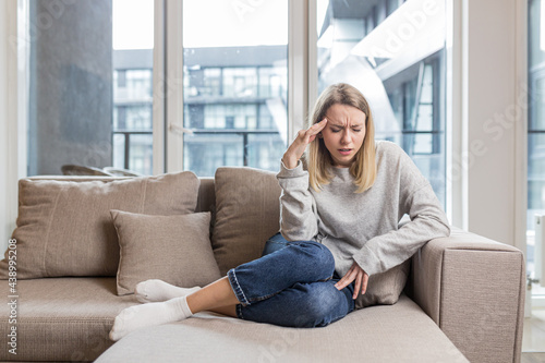 A woman holding her head anxious about an unbearable headache, sitting at home on the couch