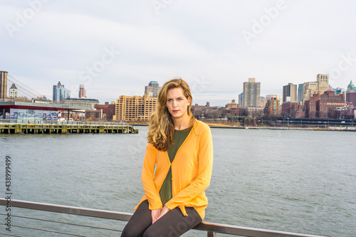 Wearing a dark yellow sweater, green underwear, a young pretty woman is sitting on a fence by a river, relaxing and thinking. Background is a big city outline..