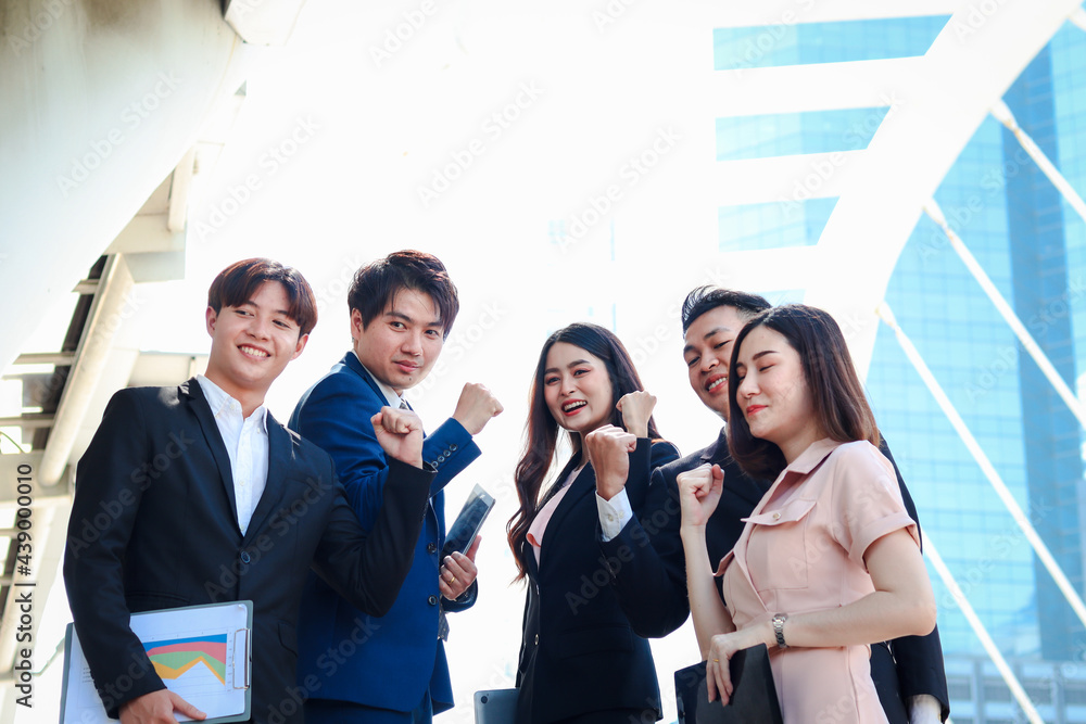 Obraz premium Group of five young Asian cheerful businesspeople raising fist hands up for celebrating success, businessman and woman cheer up their colleagues as commitment of strong teamwork at outside with skyscr