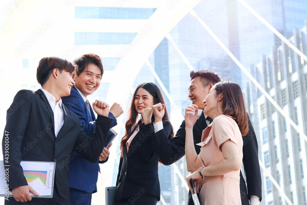 Group of five young Asian cheerful businesspeople raising fist hands up for celebrating success, businessman and woman cheer up their colleagues as commitment of strong teamwork at outside with skyscr Group of five young Asian cheerful businesspeople raising fist hands up for celebrating success, businessman and woman cheer up their colleagues as commitment of strong teamwork at outside with skyscr
