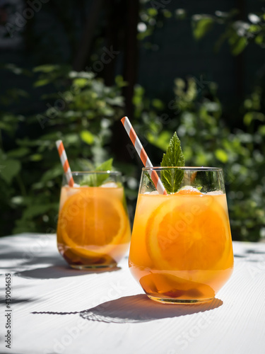 On a white wooden table in the garden, a cold orange cocktail with raspberry leaves and ice. Background - foliage from trees in the garden. Sunlight, shadow from the foliage of trees on the table.