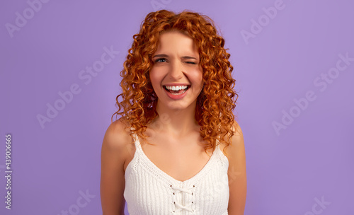 Cute redhead girl with curls winks isolated on purple background.