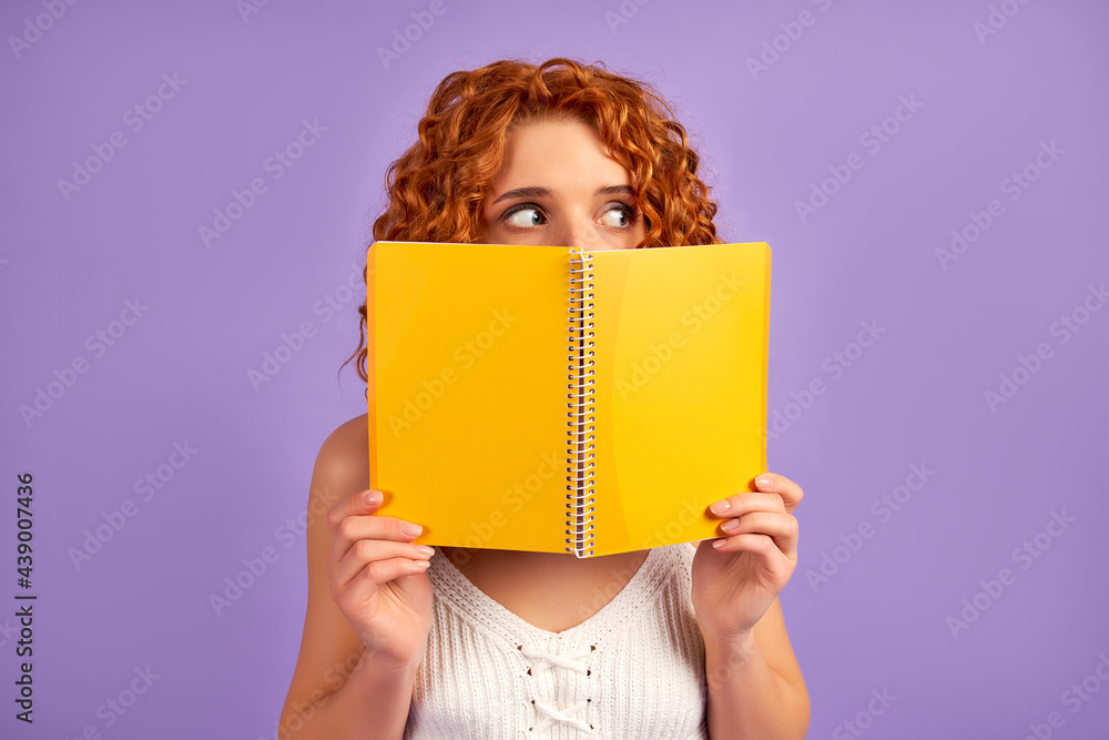 Cute redhead curly girl student teenager peeking out from behind a notebook isolated on purple background. Cute redhead curly girl student teenager peeking out from behind a notebook isolated on purple background.